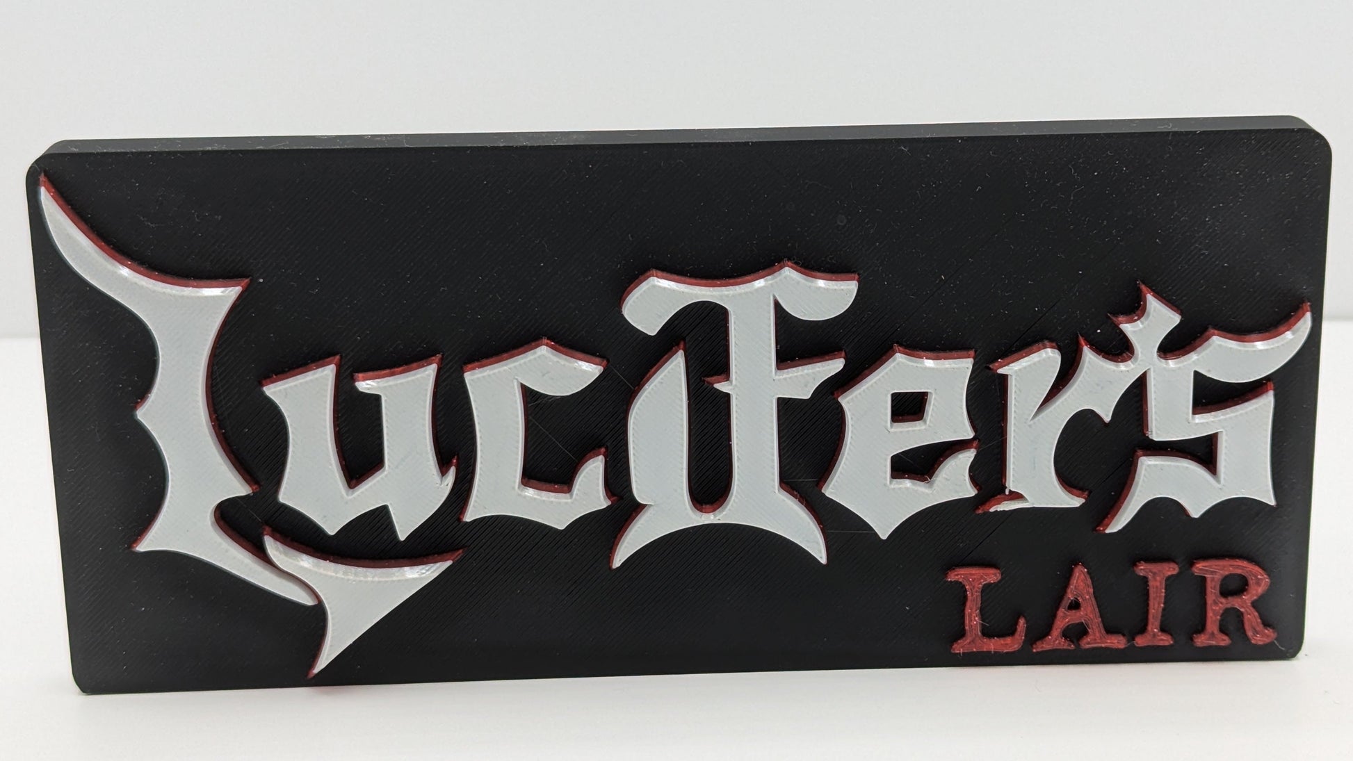 Black rectangular sign with 'Lucifer's Lair' in white and red text on a white background