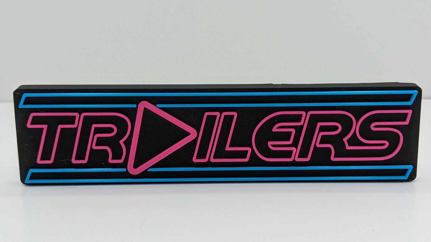 Rectangular sign with 'TRAILERS' text in pink on a black background, on a white surface.