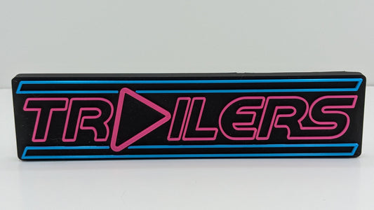 Rectangular sign with 'TRAILERS' text in pink on a black background, on a white surface.