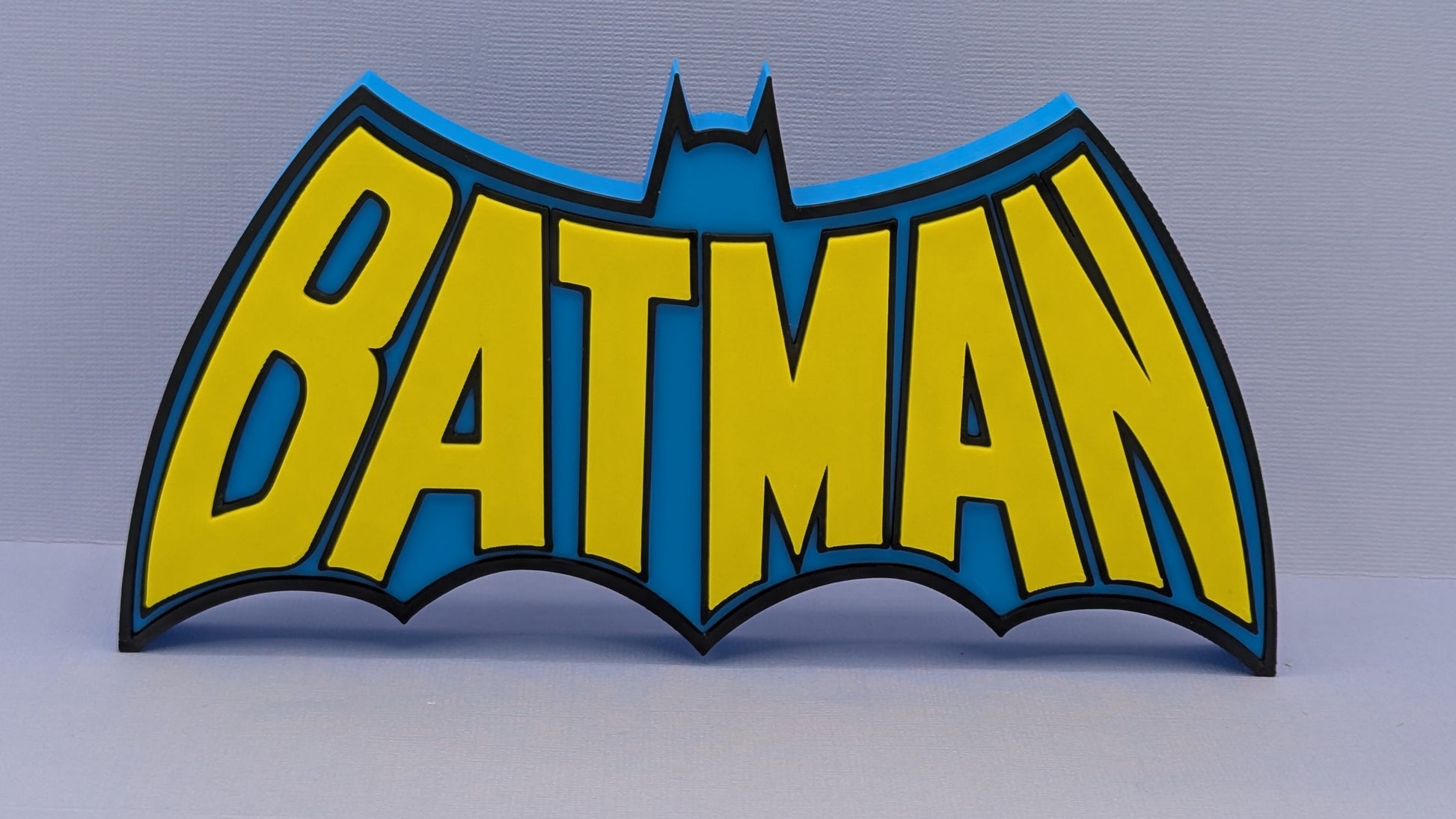 Batman logo with yellow letters and blue outline on a gray background
